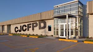 Exterior view of the CJCFPD training center building, featuring a large tan and gray modern facade with prominent “CJCFPD” lettering on the front wall. The building has a glass entrance with a multi-story window section, a paved parking area in the foreground, and clear blue skies overhead.