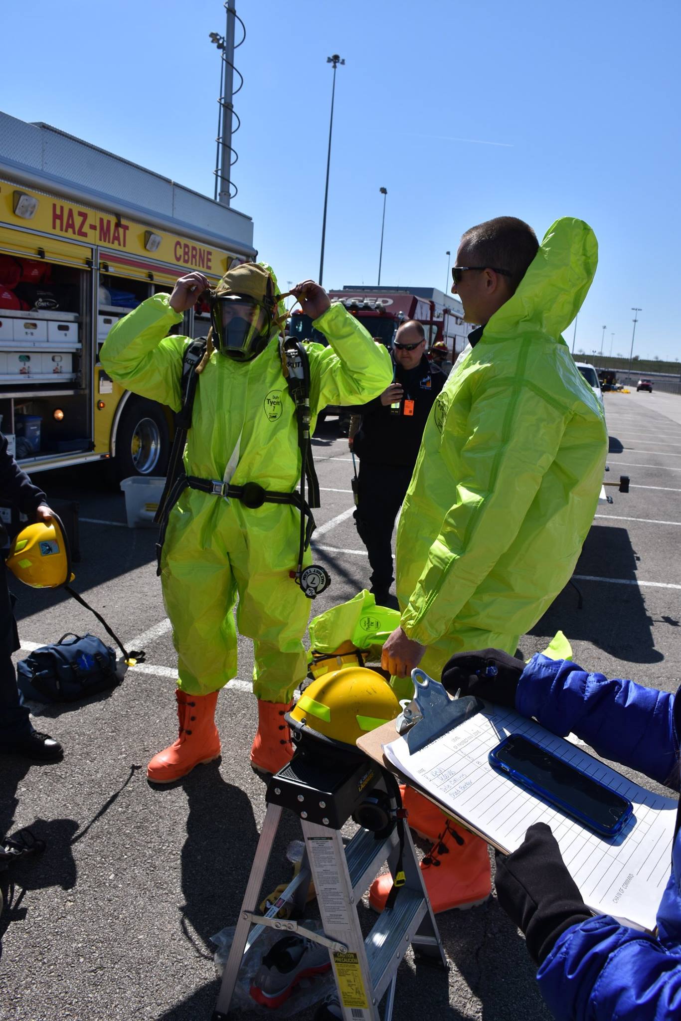 Two responders wearing bright yellow hazmat suits adjust their masks and gear beside a hazmat truck.