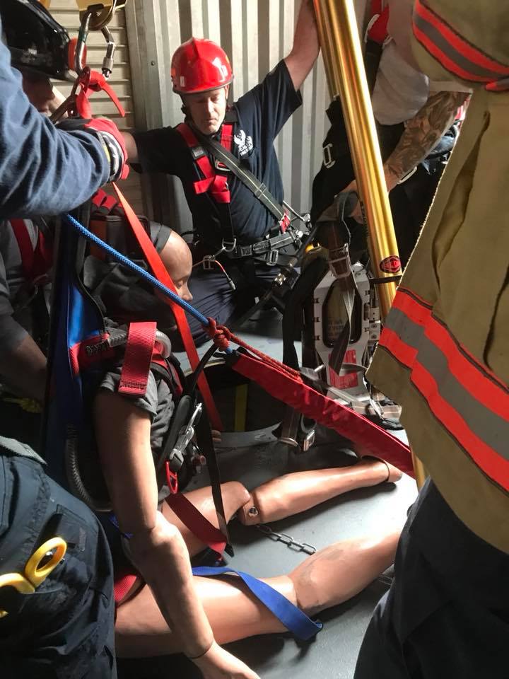 Firefighters in protective gear performing a technical rescue drill indoors, using ropes and equipment to assist a person on the ground.