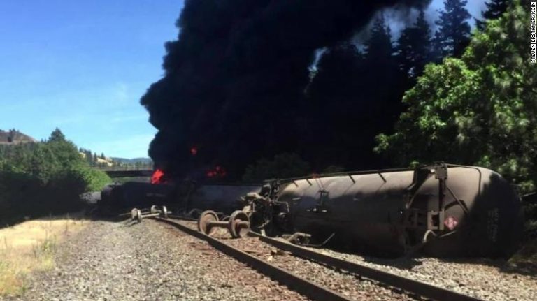 A derailed train car burns beside railroad tracks, sending up a large plume of black smoke.