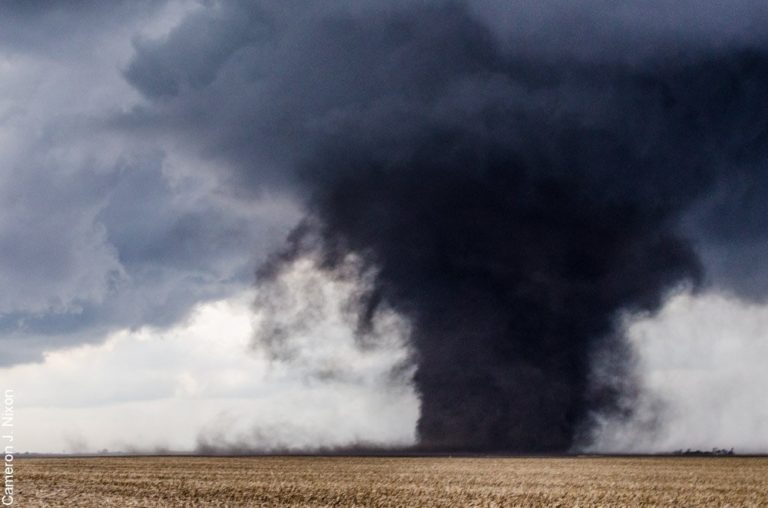 A dark tornado touches down in an open field, surrounded by swirling clouds.