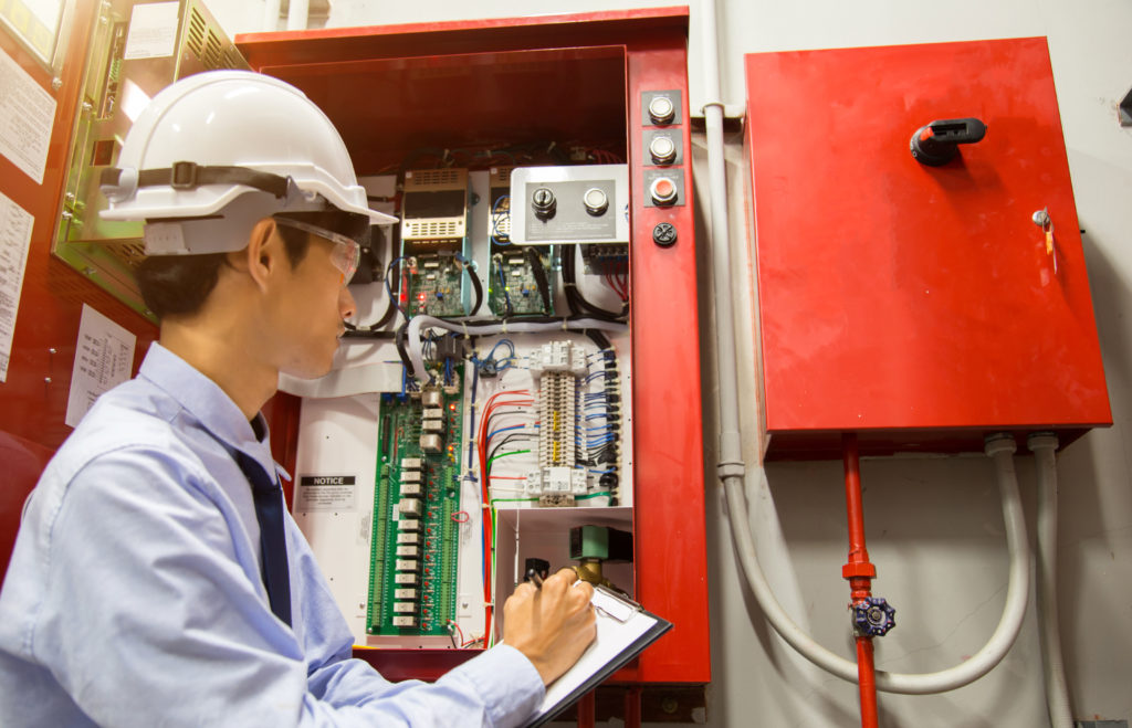 A person wearing a hard hat and safety clothing inspects an open electrical control panel filled with wiring, circuit boards, and components while writing notes on a clipboard.