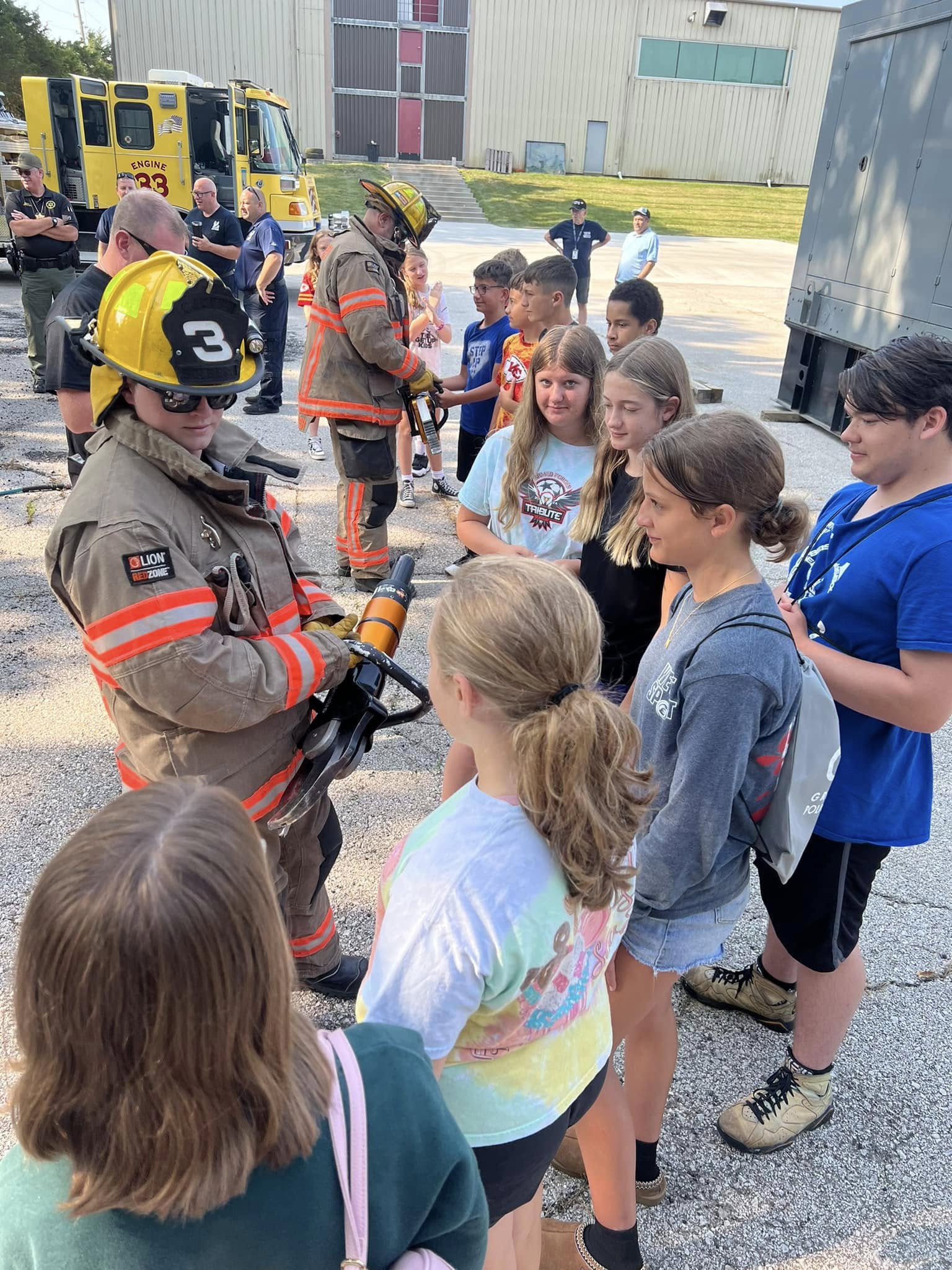 A firefighter in full gear talks to a group of elementary school students lined up outside during a safety demonstration