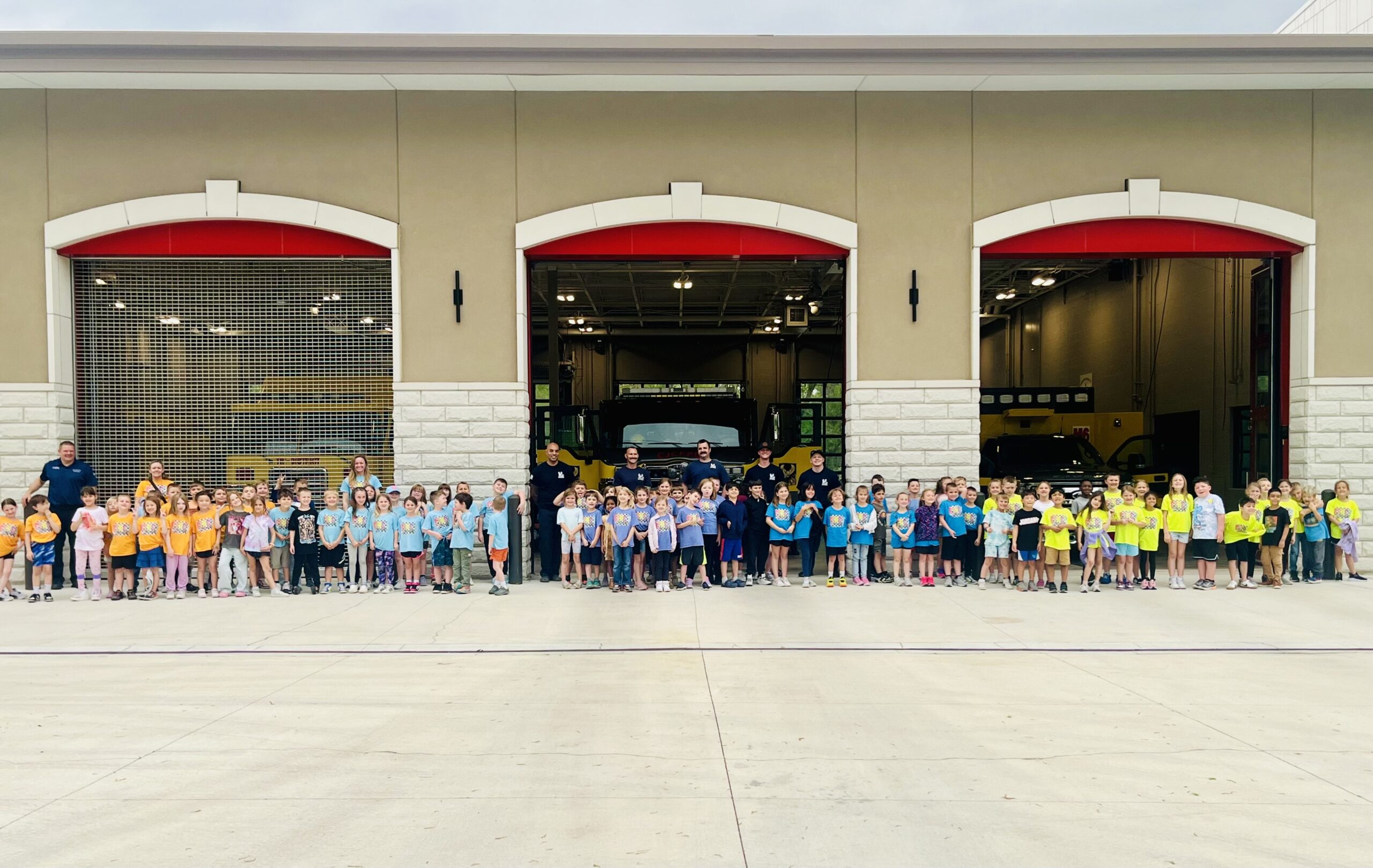 A large group of children and adults stand in front of an open fire station bay with fire apparatus visible inside