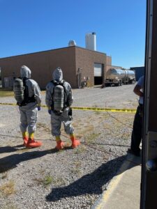 Two people in hazmat suits stand outside a commercial building near caution tape.