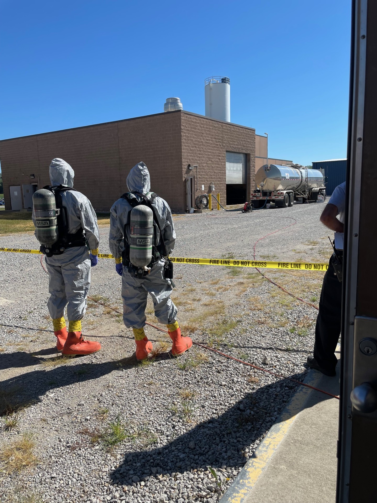 Two people in hazmat suits stand outside a commercial building near caution tape.