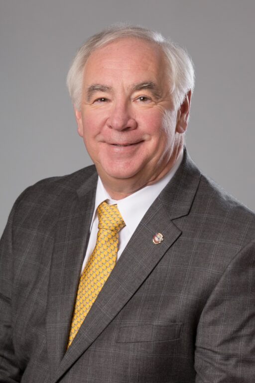 Professional head-and-shoulders portrait of a person wearing a gray plaid suit jacket, white dress shirt, and bright yellow patterned tie. The person has short white hair and is facing the camera against a plain light gray studio background. A small lapel pin is visible on the suit jacket. The image is evenly lit and framed as a formal business portrait.