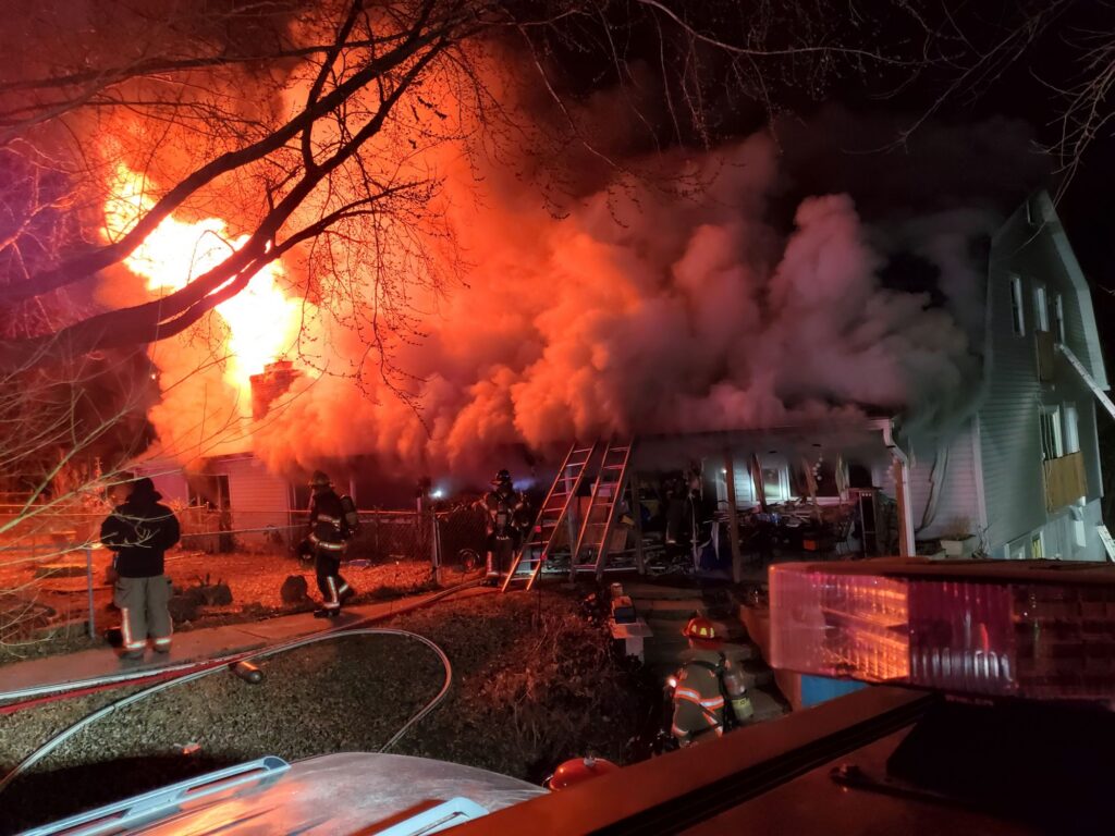 Nighttime photograph of an intense house fire with large flames erupting from the roof and heavy smoke billowing across the scene. Several firefighters are positioned around the structure, with ladders set against the building and fire hoses running across the yard and sidewalk. Emergency vehicle lights illuminate the foreground, while neighboring houses and bare tree branches frame the fire.