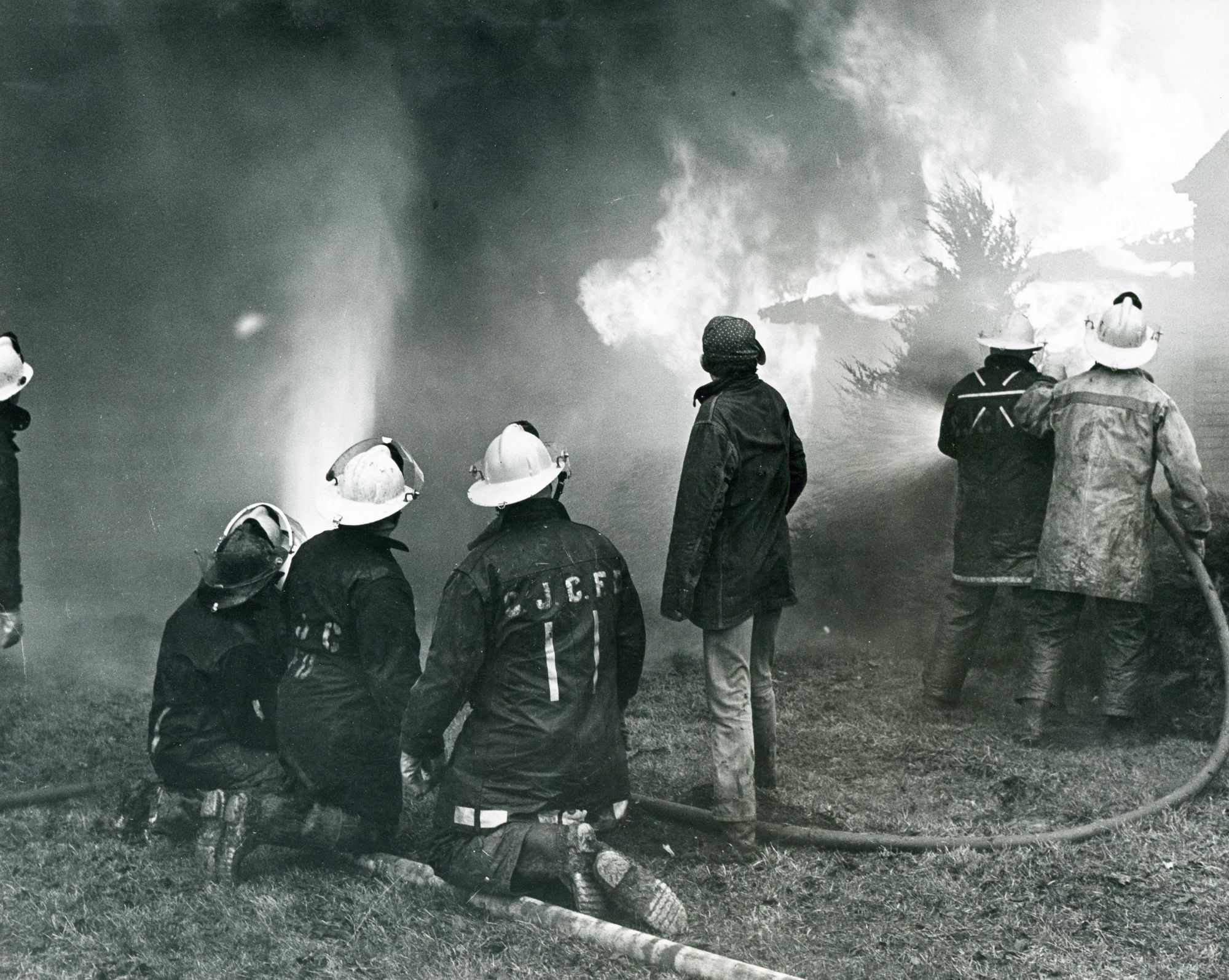 Black-and-white photograph of several firefighters battling a large structure fire. Thick smoke and flames fill the background as firefighters in helmets and protective coats kneel and stand on a grassy area, directing fire hoses toward the burning building. The image captures an active firefighting operation with heavy smoke, water spray, and flames visible throughout the scene.