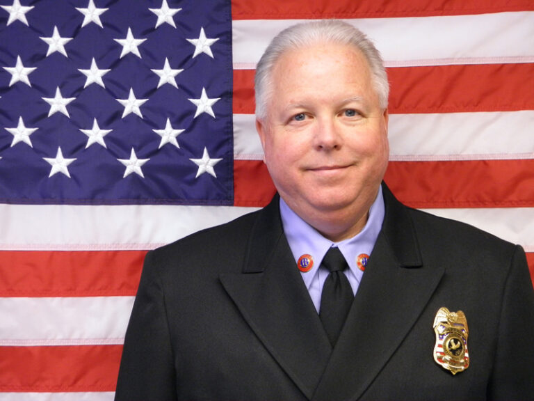 Formal portrait of a person standing in front of a large U.S. flag. The person is wearing a dark dress uniform jacket with a black tie, a light-colored shirt, collar insignia, and a badge on the left chest. The image is centered and evenly lit, with the American flag filling the background.